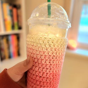 May include: A clear plastic cup with a dome lid and a green straw, filled with a blended beverage. The cup is wrapped in a handmade crochet cozy, with a gradient of pink and cream colors.