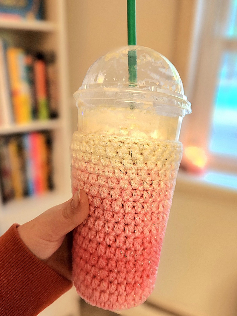 May include: A clear plastic cup with a dome lid and a green straw, filled with a blended beverage. The cup is wrapped in a handmade crochet cozy, with a gradient of pink and cream colors.