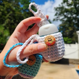 May include: Two crocheted hand sanitiser bottle holders, one white and one blue, held in a person's hand. The white holder has a wooden button that says "Handmade with Love". The blue holder has a blue strap.