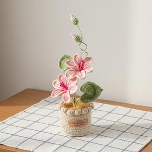 Op de afbeelding: Een handgemaakt gehaakt bloemstuk in een kleine pot. Het arrangement heeft roze bloemen met rode centra, groene bladeren en knoppen. De pot is beige met een geschulpte rand en een "HANDMADE" label.