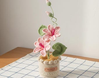 May include: A handcrafted crochet flower arrangement in a small pot. The arrangement includes pink flowers with red centres, green leaves, and buds. The pot is beige with a scalloped edge and a "HANDMADE" tag.