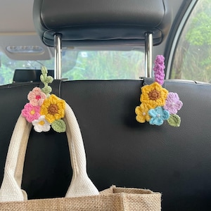 Peut inclure: Deux crochets de voiture en crochet ornés de fleurs en laine aux tons jaune, rose, bleu, blanc et violet avec des feuilles vertes. Ces accessoires décoratifs se fixent sur les tiges métalliques de l'appui-tête pour suspendre des sacs.