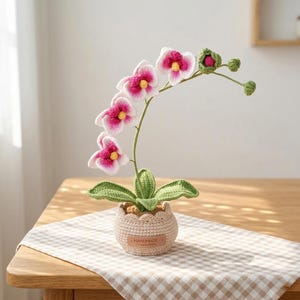 May include: A crocheted orchid plant with white and magenta petals, yellow centres, and green leaves. The plant is potted in a beige crocheted container with a brown leather-like tag that reads HANDMADE Limited edition in embossed lettering.