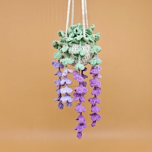 Könnte beinhalten: Ein handgefertigter, gehäkelter Hängetopf mit einem hellgrünen Oberteil, einem grauen Topf und kaskadenförmigen lila und lavendelfarbenen Blumen. Der Topf wird von weißen Kordeln vor einem warmbraunen Hintergrund gehalten.