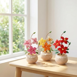 Könnte beinhalten: Drei gehäkelte Blumenarrangements in beigen Töpfen mit Spitzenbesatz. Die Blumen sind rosa, gelb und rot, mit grünen Blättern und Stielen. Die Arrangements stehen auf einem Holztisch in der Nähe eines Fensters.