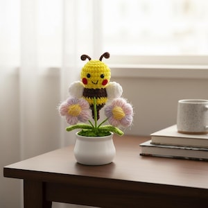 May include: A handmade crochet bee and flower arrangement in a white pot. The bee has a yellow head, brown body, and white wings. The flowers are pink and purple with yellow centers. The arrangement is on a wooden table.