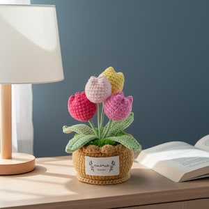 Può includere: Una composizione floreale all'uncinetto fatta a mano con cinque tulipani colorati in un vaso intrecciato. I tulipani sono in tonalità di rosa, giallo e rosa, con foglie verdi. Il vaso ha un'etichetta con la scritta "handmade".