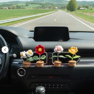 May include: Four crocheted flower decorations in small woven pots are displayed on a car dashboard. The flowers are white, red, pink, and yellow, with green leaves. The car interior is black, and a road is visible in the background.