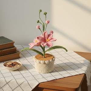 May include: A crocheted flower arrangement featuring pink flowers with yellow centres and green leaves, in a white pot with a scalloped edge. The pot sits on a checkered tablecloth. A bowl of buttons and books are in the background.