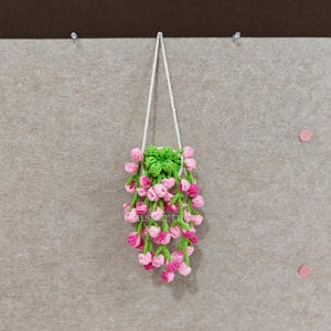 May include: A handmade crochet floral hanging decoration. The piece features a green base with pink and white flowers cascading downwards. The flowers are attached to green stems. A white cord is used for hanging.