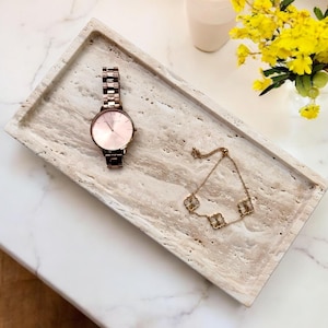 Puede incluir: Una bandeja rectangular de travertino beige con un reloj de oro rosa y una pulsera dorada con abalorios en forma de trébol. El reloj tiene una esfera rosa y una correa de metal. Flores amarillas en un jarrón en el fondo.