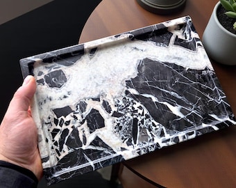 Op de afbeelding: Een rechthoekige decoratieve tray met een zwart-wit marmerpatroon. De tray wordt door een hand vastgehouden. De tray heeft een gepolijste afwerking en is geschikt voor woondecoratie.