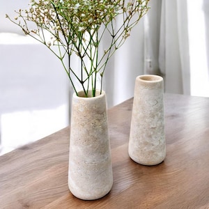 Op de afbeelding: Twee beige stenen vazen met een textuuroppervlak staan op een houten tafel. Eén vaas bevat delicate witte bloemen met dunne groene stelen. De vazen hebben een taps toelopende vorm, breder aan de basis en smaller aan de bovenkant.