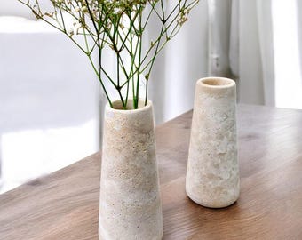 Puede incluir: Dos jarrones de piedra beige con superficie texturizada sobre una mesa de madera. Un jarrón contiene delicadas flores blancas con finos tallos verdes. Los jarrones tienen forma cónica, más anchos en la base y más estrechos en la parte superior.