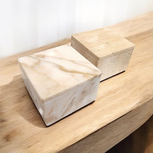 May include: Two decorative boxes with lids. One box is a light beige stone with a speckled texture. The other box is a triangular shape with a white and beige marble pattern. Both boxes are on a wooden surface.