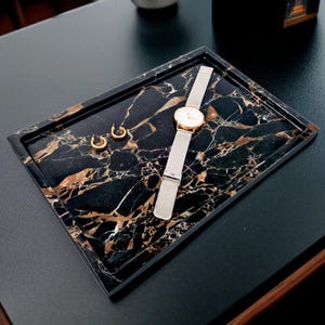 May include: A rectangular black marble tray with gold veining, holding a gold watch with a silver band and two gold hoop earrings. The tray is on a dark surface, showcasing the jewelry and watch.