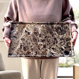 Puede incluir: Una bandeja rectangular con un patrón similar al mármol en tonos marrón, negro y crema. La bandeja está sostenida por una persona. Las dimensiones de la bandeja son de aproximadamente 46 cm de largo y 30 cm de ancho.
