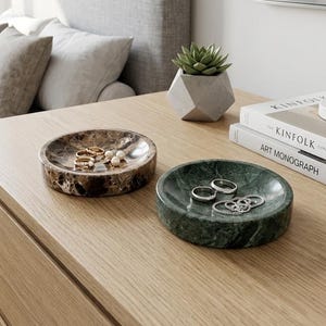 Peut inclure: Deux vide-poches ronds en marbre, un marron et un vert, posés sur une surface en bois. Le plat marron contient des bagues et des perles, tandis que le plat vert expose des bagues en argent. Une plante succulente et des livres sont à proximité.