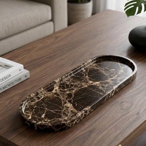 May include: An oval-shaped tray with a dark brown marble pattern rests on a dark wood coffee table. The tray has a raised edge. A black vase with green leaves and two books are also on the table. The table has a lower shelf and gold-coloured legs.