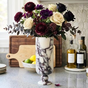 Puede incluir: Un jarrón cilíndrico de mármol con vetas blancas y moradas oscuras, con una base de pedestal. El jarrón contiene un arreglo floral de rosas color crema, ranúnculos morados oscuros, anémonas violetas y follaje de eucalipto verde.