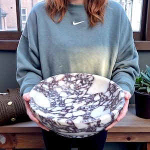 Könnte beinhalten: Eine große, runde Marmorschale mit einem weißen und dunkelviolett geäderten Muster. Die Schale wird von einer Person gehalten. Ein brauner Pullover und eine Topfpflanze befinden sich im Hintergrund.