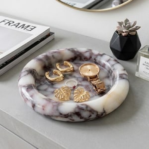 May include: A marble jewellery tray with gold earrings, a ring, and a watch. The tray has a circular shape with a white and purple marbled pattern. A small succulent plant in a black geometric pot and a perfume bottle are also present.