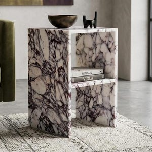 May include: A marble side table with a white and purple veined pattern. The table has a shelf with two books titled "Minimal" and "Viola". A dark metal bowl and a black cat sculpture sit on top. The table is 50 cm wide.