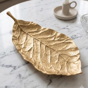 Peut inclure: Un plateau décoratif doré en forme de grande feuille avec une surface texturée et une nervure centrale saillante. Le plateau présente une extrémité pointue et une petite tige à la base, arborant une finition métallique aux détails organiques.
