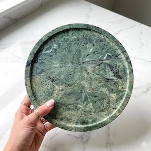 Peut inclure: Un plateau circulaire en marbre vert doté d'une surface polie avec des veines sombres naturelles et un rebord surélevé. La pierre présente un motif varié de vert forêt profond, de sauge et de stries noires sur toute la structure circulaire.