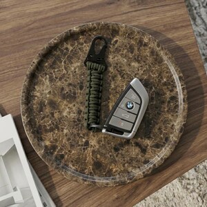 Puede incluir: Una llave de coche y un llavero de paracord verde descansan sobre una bandeja de m&aacute;rmol marr&oacute;n. La llave tiene un logotipo de BMW. La bandeja est&aacute; sobre una superficie de madera oscura, con una revista visible.