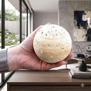 May include: A spherical object made of beige, porous travertine stone with a natural, textured surface. The stone sphere features visible pits and organic striations throughout its round form, showcasing the raw, earthy characteristics of the material.