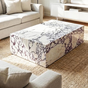 Könnte beinhalten: Ein rechteckiger Couchtisch aus Marmor mit einem weißen und violett geäderten Muster. Der Tisch ist ein massiver Block mit polierter Oberfläche. Der Tisch steht auf einem gewebten Jute-Teppich, mit einem hellen Sofa im Hintergrund.