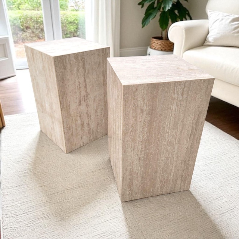 May include: Two beige, rectangular pedestals with a textured, stone-like appearance. The pedestals are placed on a light-colored rug, and are in front of a window and a white couch.