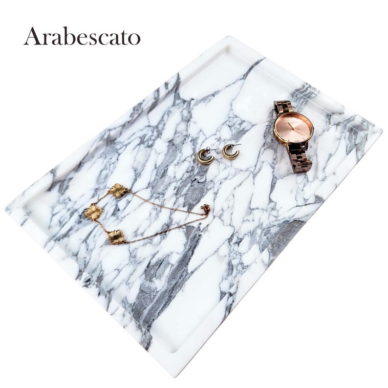 Puede incluir: Una bandeja rectangular con dise&ntilde;o de m&aacute;rmol, con base blanca y vetas grises. La bandeja contiene un reloj dorado, pendientes y un collar. La palabra "Arabescato" est&aacute; impresa en la esquina superior izquierda. Ideal para exhibir joyas.