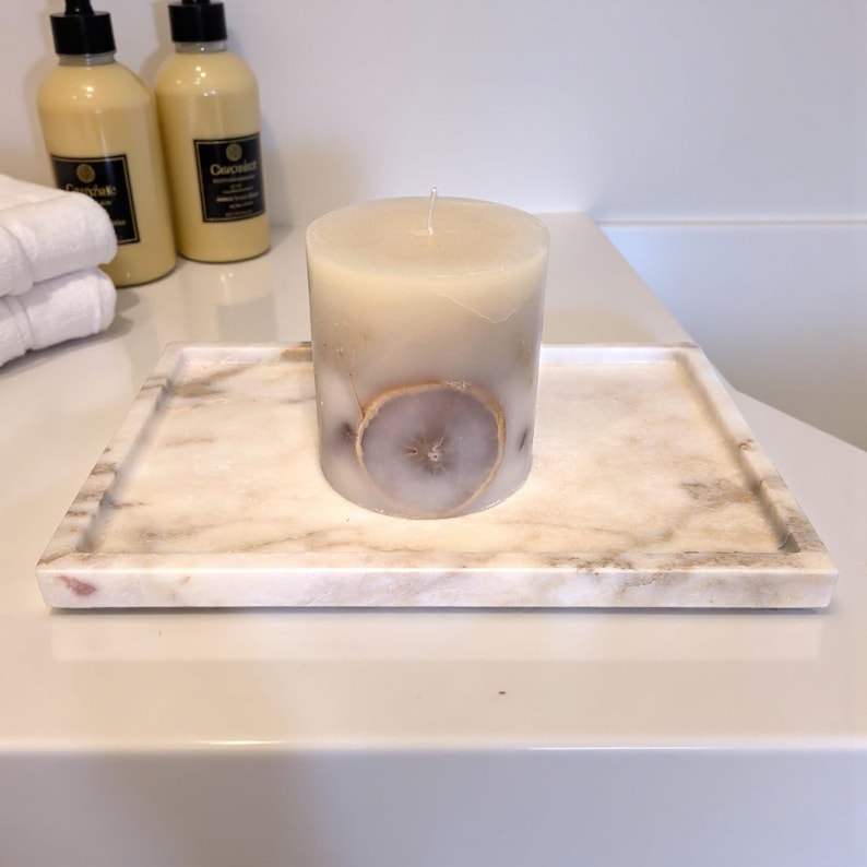 May include: A cream-colored pillar candle with a dried citrus slice embedded in the wax, resting on a rectangular marble tray. The tray has a light beige and pink veining pattern. The candle and tray are on a white surface.