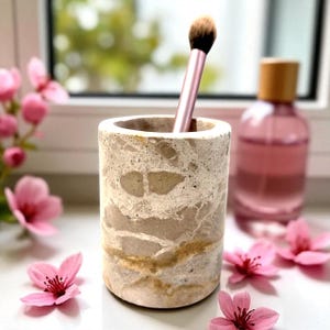 May include: A cylindrical, beige marble makeup brush holder with a pink brush inside. The holder has a marbled pattern with brown and tan veins. A pink perfume bottle and pink flowers are in the background.