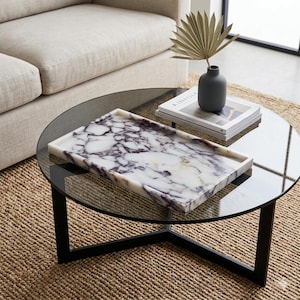 May include: A rectangular marble tray with a white and purple veined pattern rests on a round glass coffee table with a black metal frame. A small grey vase with a dried palm leaf and a book are also on the table.