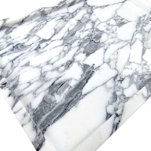 Puede incluir: Una bandeja rectangular con dise&ntilde;o de m&aacute;rmol. La bandeja tiene una base blanca con vetas grises y negras. Los bordes est&aacute;n ligeramente elevados, creando un borde. Fabricada con un material s&oacute;lido, ideal para servir.