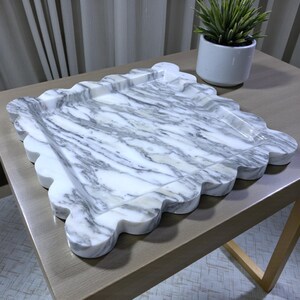 Könnte beinhalten: Ein dekoratives Marmortablett mit gewelltem Rand. Das Tablett hat ein weißes und graues Adernmuster. Eine kleine Topfpflanze steht im Hintergrund auf einem Holztisch.
