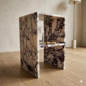 May include: A marble side table with a shelf. The table is white with dark purple veining. A clear glass bottle and a gold spoon rest on the shelf. The table is on a wooden floor.