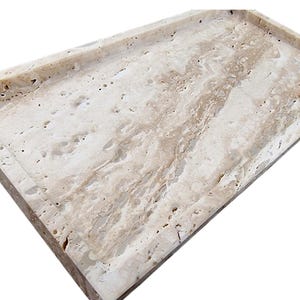 May include: Rectangular travertine tray with a raised border. The tray features a natural beige and cream color palette with unique veining patterns. Ideal for serving or decorative use.