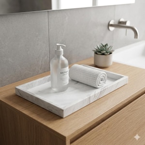 May include: A bathroom countertop arrangement featuring a marble tray with a rolled white towel, a clear soap dispenser with the text "MERA", and a small potted succulent. The countertop is made of light wood.