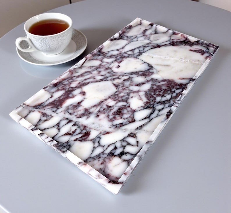 May include: A rectangular marble tray with a purple and white pattern. A white teacup and saucer with tea are on the left. The tray is a decorative item for serving or displaying items.