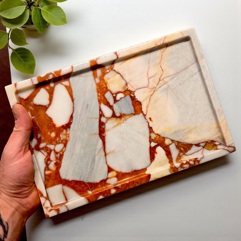 May include: Rectangular marble tray with a unique pattern of white, gray, and orange-red veining. The tray has a raised edge and is held by a hand. The tray is a decorative item.