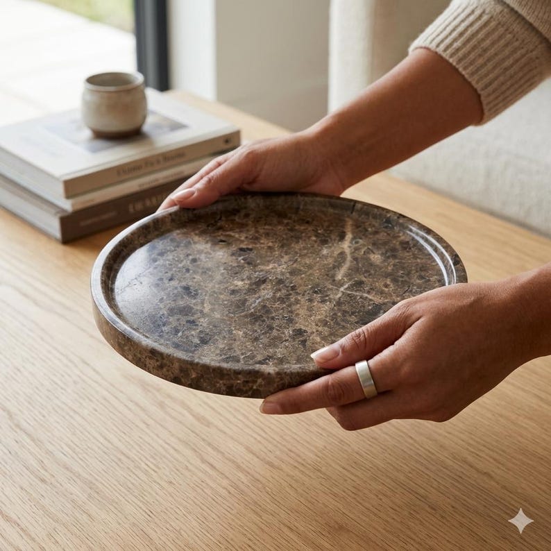 May include: A circular, brown marble tray with a raised edge. The tray is held by two hands. The marble has a natural, veined pattern. The tray is on a wooden surface.