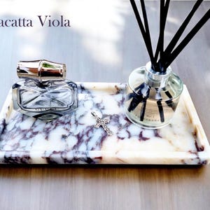 May include: A rectangular marble tray with a perfume bottle, a silver cross pendant, and a reed diffuser. The tray has a white and purple veined pattern. The perfume bottle is clear with a silver cap. The reed diffuser has black reeds.