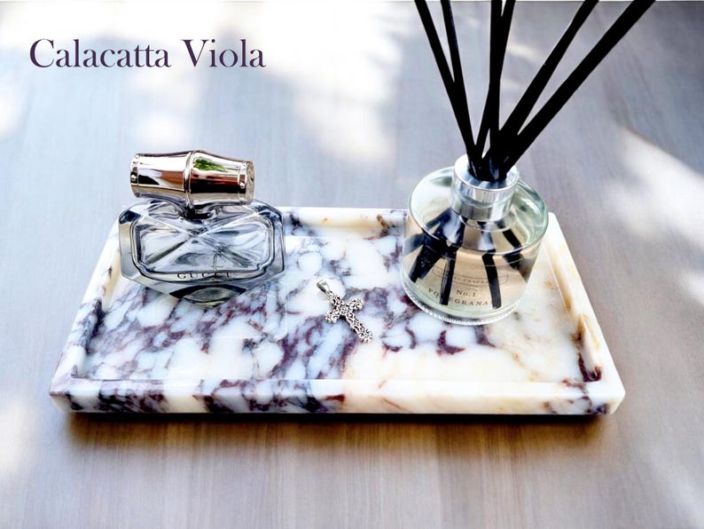 May include: A rectangular marble tray with a perfume bottle, a silver cross pendant, and a reed diffuser. The tray has a white and purple veined pattern. The perfume bottle is clear with a silver cap. The reed diffuser has black reeds.