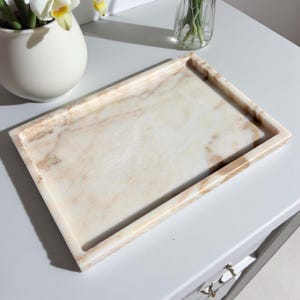 May include: A rectangular marble tray with a light beige and brown veined pattern. The tray has a raised edge and is sitting on a white surface. The tray is empty and ready to be used for decorative purposes.