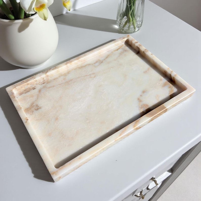 May include: A rectangular marble tray with a light beige and brown veined pattern. The tray has a raised edge and is sitting on a white surface. The tray is empty and ready to be used for decorative purposes.