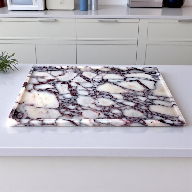 May include: Rectangular marble tray with a white base and intricate purple and black veining. The tray has a slightly raised edge. It is set on a white countertop. Suitable for serving or display.
